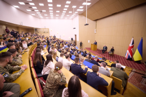 Eine große Gruppe von Menschen an Tischen in einem Raum mit Flaggen auf der rechten Seite, eine Wand im Hintergrund, Papiere auf den Tischen, einigestehend in der Nähe eines Podiums mit einem Mikrofon und Deckenleuchten und ein Projektor.