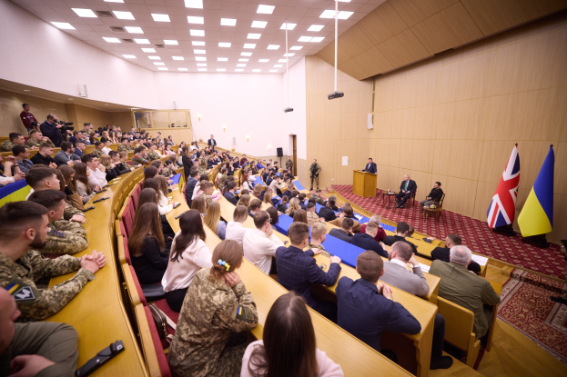 Eine große Gruppe von Menschen an Tischen in einem Raum mit Flaggen auf der rechten Seite, eine Wand im Hintergrund, Papiere auf den Tischen, einigestehend in der Nähe eines Podiums mit einem Mikrofon und Deckenleuchten und ein Projektor.
