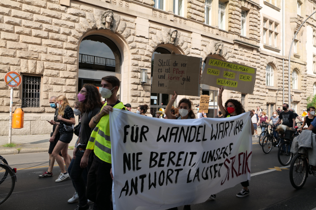 Eine Gruppe von Menschen marschiert auf der Straße in Berlin, hält Schilder und Banner hoch und fährt mit Fahrrädern, vor einem Gebäude mit Fenstern, Bögen, Säulen, Skulpturen, Bäumen und einem Laternenmast, an der Protestaktion gegen die Anti-Kriegsbewegung teilnehmend.