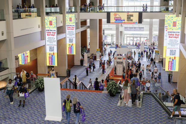 Eine große Gruppe von Menschen, die durch ein großes Gebäude mit Bannern, Topfpflanzen, Rolltreppen, Geländern, Säulen, bedruckten Schildern, Glastüren und anderen Gegenständen geht. Einige tragen Taschen, wahrscheinlich nehmen sie am Nationalen Buchfestival teil.