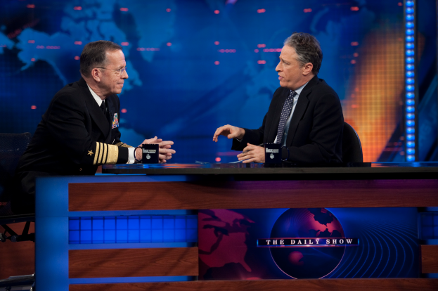 Zwei Männer in schwarzen Blazern, Hemden und Krawatten sitzen an einem Tisch mit Mikrofonen dazwischen und unterhalten sich vor einem Fernseher und einem blauen Hintergrund.