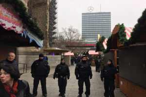 Eine Gruppe von Polizisten in schwarzen Uniformen und Mötzen steht vor einem geschmückten Weihnachtsmarkt in Berlin, mit Gebäuden, Bäumen und einem klaren blauen Himmel im Hintergrund und einer Tafel mit Text links sichtbar.
