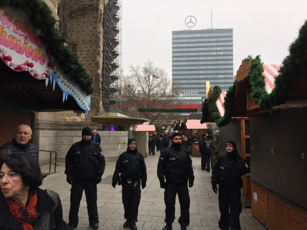 Eine Gruppe von Polizisten in schwarzen Uniformen und Mötzen steht vor einem geschmückten Weihnachtsmarkt in Berlin, mit Gebäuden, Bäumen und einem klaren blauen Himmel im Hintergrund und einer Tafel mit Text links sichtbar.