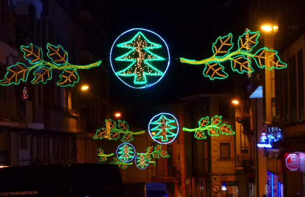 Eine städtische Straße, die mit festlichen Weihnachtslichtern geschmückt ist, die Blätter und andere Schmuckstücke zeigen, mit Fahrzeugen, die auf beiden Seiten an Gebäuden vorbeifahren.