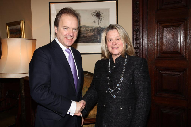 Eine Frau und ein Mann in einem Anzug lächeln, schütteln sich die Hände und sehen sich an, mit einer Wand, Fotorahmen, einer Lampe, einer Tür und ein paar Gegenständen im Hintergrund.