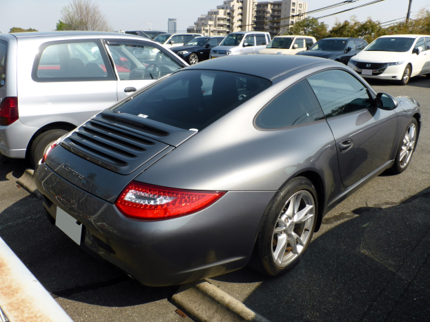 Ein silberner Porsche 911 auf einem Parkplatz mit anderen Autos, Gebäuden, Bäumen, Masten, Drähten und einem klaren blauen Himmel.
