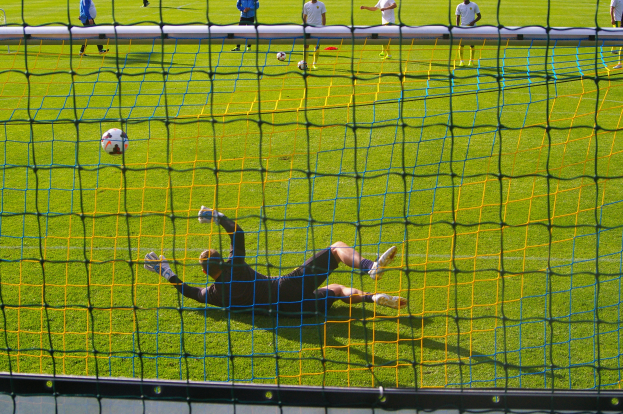 Ein Fußballspieler hält den Ball vor dem Tor, mit einem Netz im Vordergrund und einigen Menschen im Hintergrund, sowie mehreren Bällen auf dem Boden verteilt.