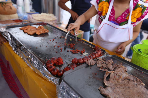 Eine Frau in einer Schürze kocht auf einem Grill auf einem Markt, mit einem Tisch voller Lebensmittel, einem Eimer und anderen Gegenständen in der Nähe und ein paar Menschen im Hintergrund.