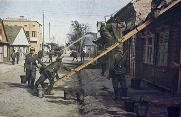 Deutsche Soldaten im Zweiten Weltkrieg in Deutschland stehen auf einer Straße neben einem Gebäude, tragen Helme und halten hölzerne Gegenstände, mit Eimern auf dem Boden und Gebäuden, Bäumen, Polen und Drähten im Hintergrund unter einem klaren Himmel.