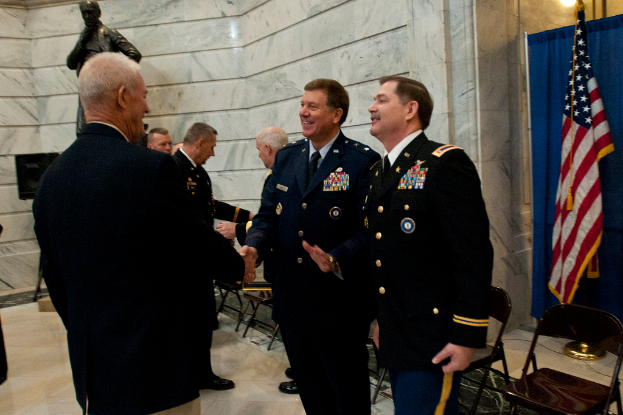 Menschen geben sich die Hand vor Stühlen, mit einer Statue, einer Flagge und einem Vorhang im Hintergrund.