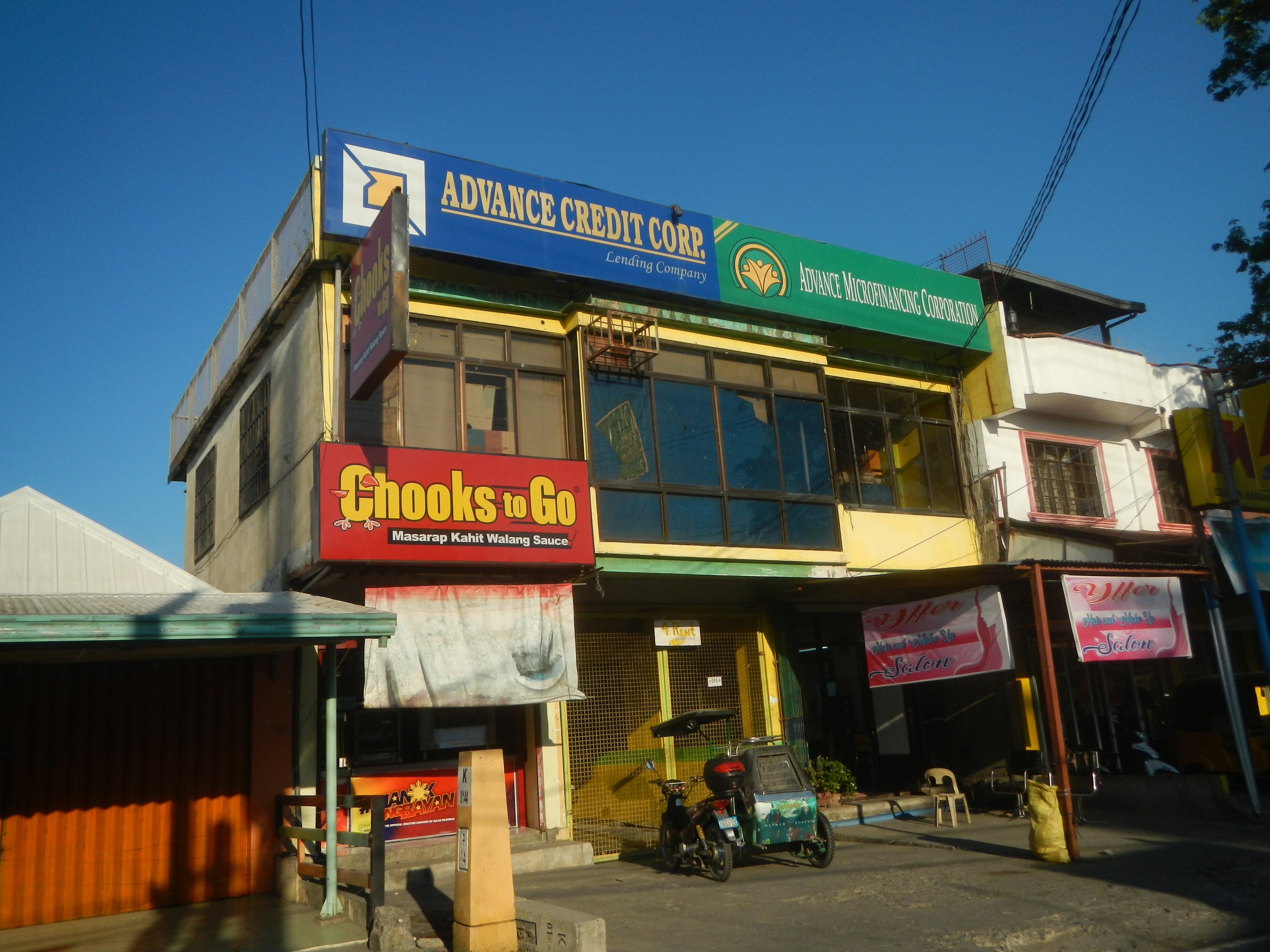 Ein Geschäftsgebäude mit einer Tafel, auf der "Advance Credit Corp" steht, an einer Straße mit Fahrzeugen, Strommasten und Bäumen unter einem klaren Himmel.