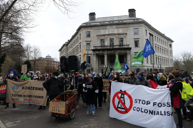 Eine große Gruppe von Menschen marschiert in einem Protest gegen fossile Brennstoffe, trägt Transparente und Fahnen, mit einem Fahrzeug im Vordergrund und Gebäuden, Bäumen und einem klaren blauen Himmel im Hintergrund.