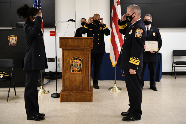 Eine Gruppe uniformierter Personen in Masken steht vor einem Podium mit einem Mikrofon, umgeben von Fahnen, Stühlen und Tischen, während einer Vereidigung für einen neuen Feuerwehrchef, wobei eine Person ein Zertifikat hält.