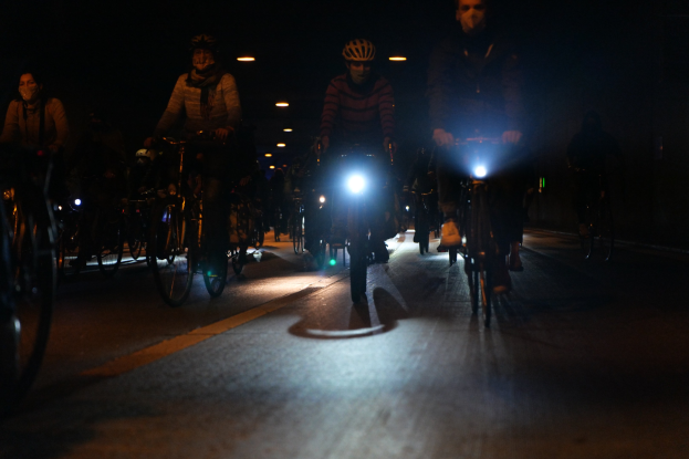 Eine Gruppe von Menschen, die nachts auf Fahrrädern durch eine gut beleuchtete Stadtstraße fahren, alle tragen Helme.