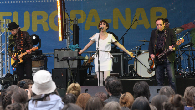 Eine Gruppe von vier Musikern, die auf der Bühne eines Konzerts spielen, mit einer singenden Frau in der Mitte, zwei Gitarristen zu beiden Seiten, einer Menge im Vordergrund und Lautsprechern im Hintergrund.