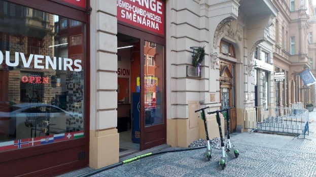 Eine stadtische Straße mit parkenden Rollern, Gebäuden, Texttafeln, einem Geländer, Bäumen und einem klaren blauen Himmel.