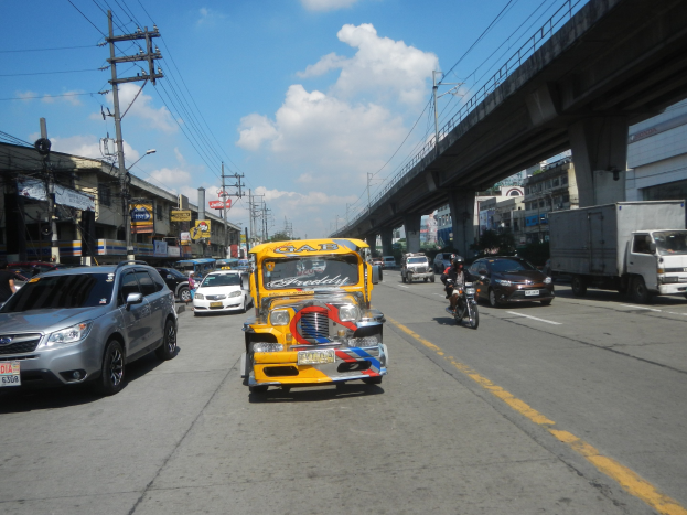 Gelbes Taxi fährt auf einer Stadtstraße mit hohen Gebäuden, Stromleitungen und einer Brücke im Hintergrund unter einem bewölkten Himmel, mit einem Wasserzeichen auf dem Bild.