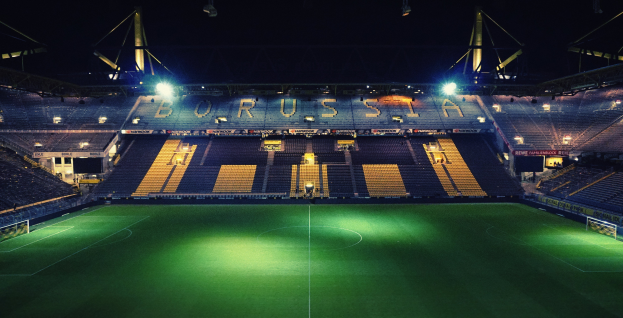 Ein Fußballfeld bei Nacht mit hellen Stadionlichtern, die den saftig grünen Rasen beleuchten, und einigen Menschen auf den Tribünen.