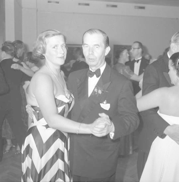 Ein Mann in einem schwarzen Blazer, weißem Hemd und schwarzen Hosen tanzt mit einer Frau in unbekannter Kleidung auf einer formellen Veranstaltung, indem sie sich an den Händen halten und zur Musik an einer Wand schwenken.