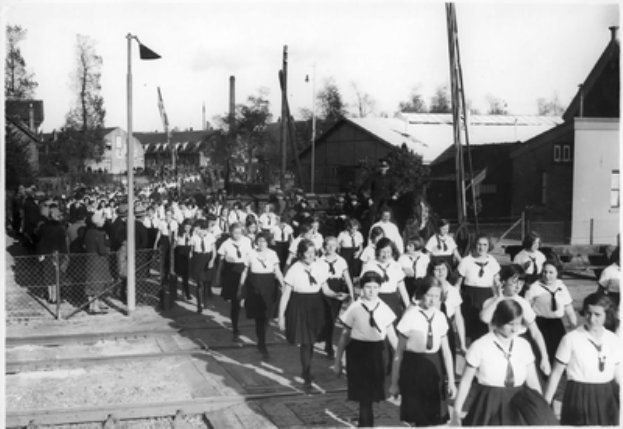 Eine Gruppe von Mädchen in Schuluniformen marschiert auf einer Straße, mit einem Zaun auf der linken Seite und Menschen, die dahinter stehen, Polen, Bäume und Gebäude im Hintergrund, unter einem sichtbaren Himmel, in Schwarz-Weiß.