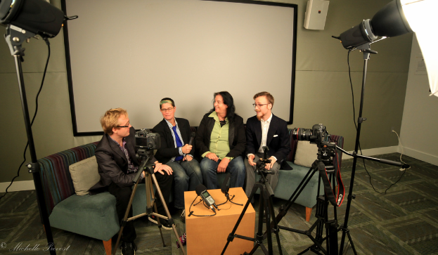 Eine Gruppe von drei Personen sitzt auf einem Sofa und gibt ein Interview. Links ist eine Person in Schwarz, in der Mitte eine Person mit blauer Krawatte und Mikrofon und rechts eine Person, die strahlt. Hinter ihnen ist ein Projektionsschirm und vor ihnen ein Tisch mit Mikrofonaufnahmegerät, Kameras und großen Scheinwerfern.