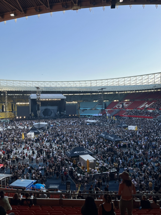 Große Menge sitzend und stehend in einer Halle mit Zelten, Geländern und Deckenbeleuchtung vor einem sichtbaren Himmel während eines Konzerts in der O2 Arena in London.