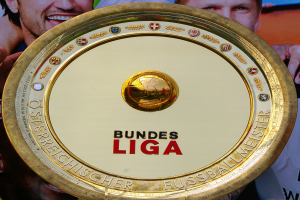 Ein Mann hält ein goldenes Schild mit der Aufschrift "Bundesliga" hoch, im Hintergrund ist eine Fahne mit drei Personen zu sehen.