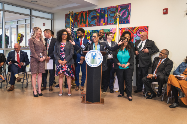 Eine Gruppe von Menschen, die vor einem Rednerpult mit Mikrofon und einer Tafel mit der Aufschrift "Sancho City Councilman Joins City Council" stehen und sitzen, mit Flaggen, Feuerlöscher und Deckenlampen an der Rückwand.