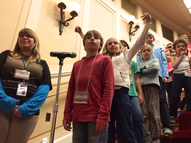 Kinder singen vor einem Mikrofon mit einer Frau neben ihnen, Stühle rechts und beleuchtete Wand im Hintergrund.
