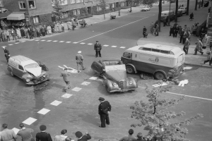 Schwarz-weiß-Szene eines Autounfalls an einer Straßenkreuzung mit mehreren Fahrzeugen, darunter ein Lieferwagen, und einer Gruppe von Zuschauern; Gebäude, Bäume, Pfosten und Texttafeln sind im Hintergrund sichtbar.