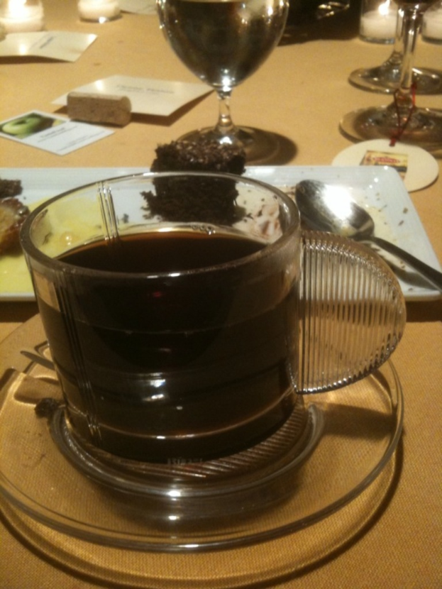 Ein Tisch mit Tasse, Untertasse, Tee, Teller, Löffel, Brownie, Glas, Wein, Kerze und zusätzlichem Essen.