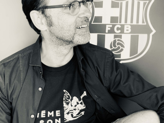 Ein Mann mit Brille und schwarzem Hemd lächelt vor einer Wand mit dem FC Barcelona-Logo, aufgenommen in Schwarz-Weiß.