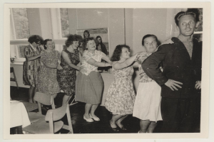 Eine Gruppe von Menschen, die in einem Raum mit Stühlen und einem mit einer wei├čen Decke bedeckten Tisch tanzen. An der Wand h├Ąngt ein Photo-Rahmen und im Hintergrund sind Fenster zu sehen.