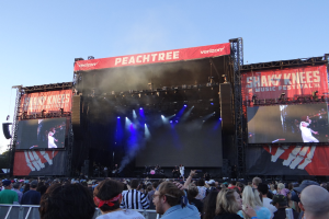 Eine große Menschenmenge steht vor einer hell erleuchteten Bühne auf dem Shaky Knees Music Festival, auf der Musiker hinter einem Geländer unter einem klaren blauen Himmel spielen.