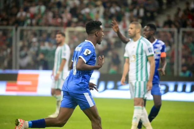Ein Fußballspieler in einem blauen und weißen Trikot feiert ein Tor, umgeben von Teamkollegen in weißen und grünen Trikots, vor einer jubelnden Menge.