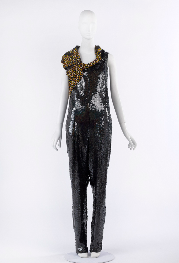 Ein Schaufensterpuppen-Mann in einem schwarzen und goldenen Pailletten-Jumpsuit mit einem Leopardenmuster-Schal um den Hals, die Pailletten glitzern und reflektieren das Licht.