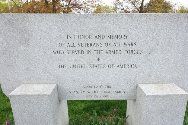 Ein Gedenkstein mit einer Inschrift für alle U.S.-Armed-Forces-Veteranen, umgeben von Blumen, Gras, Bäumen und einem bewölkten Himmel.