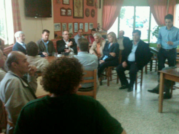 Eine Gruppe von Menschen, die in einem Raum auf Stühlen sitzen, mit einer Person in einem blauen Hemd, die steht, einem Fernseher, Gemälden an der Wand, einem Glasfenster, durch das man Fahrzeuge und Bäume sehen kann, und Vorhängen.