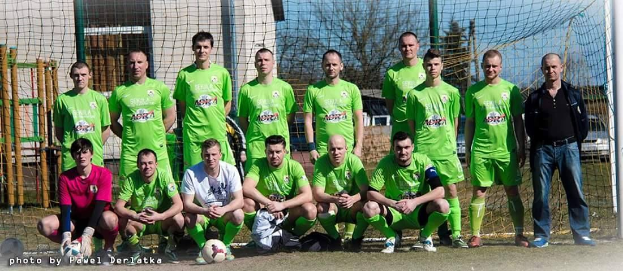 Gruppe von Männern in grünen T-Shirts auf einem Fussballfeld mit einem Ball und einem Torpfosten, Bäume und Gebäude im Hintergrund.