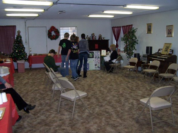 Ein Raum mit mehreren Stühlen, Tischen und Menschen, einige stehen, andere sitzen, mit Tassen und Gefäßen auf den Tischen, einem Weihnachtsbaum, einem Piano auf der rechten Seite, Rahmen, Fenstern und Röhrenleuchten an der Decke und einer Wand mit einem Strauß, einem Vorhang, einer Tür und weiteren Rahmen im Hintergrund.