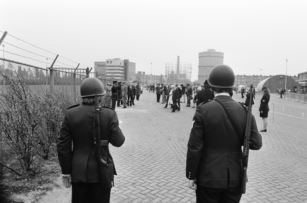 Zwei Polizeibeamte in Helmen und mit Waffen in den Händen stehen nebeneinander auf einem Bürgersteig, links davon ein Zaun und Pflanzen, Gebäude und Masten im Hintergrund und ein klarer Himmel.