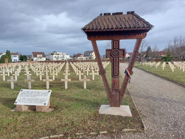 Ein Friedhof in Stuttgart, Deutschland, mit einem Pavillon in der Mitte, umgeben von Gras, einem Schild mit Text, Bäumen, Gebäuden und einem bewölkten Himmel, der an die Opfer des Zweiten Weltkriegs erinnert.