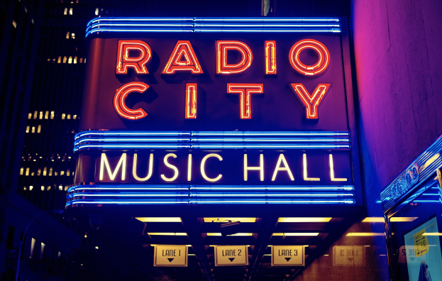 Außenansicht des Radio City Music Hall in New York City bei Nacht, hell erleuchtet und von hohen Gebäuden flankiert, mit einem "Radio City"-Schild und mehreren kleineren Schildern auf der rechten Seite.
