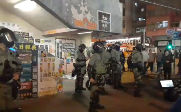 Polizeibeamte in Schutzausrüstung stehen vor einer Menge von Helmträgern mit Schildern, mit Plakaten an den Wänden, Deckenleuchten und Gebäuden im Hintergrund in Hong Kong.