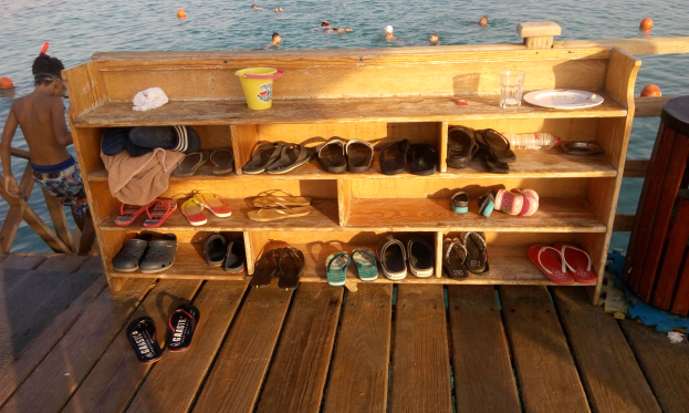 Eine hölzerne Bank auf einem Dock mit verschiedenen Schuhen, einem Becher, einer Flasche, einem Teller, einem Glas und anderen Gegenständen, daneben ein Mülleimer und im Hintergrund schwimmende Menschen.
