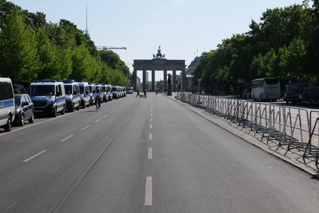 Eine Reihe von Polizeiwagen, die auf einer Straße vor dem Brandenburger Tor in Berlin, Deutschland, geparkt sind, mit Menschen auf Fahrrädern und in der Nähe stehenden, Barrieren, Bäumen und einem Bogen mit Statuen im Hintergrund.