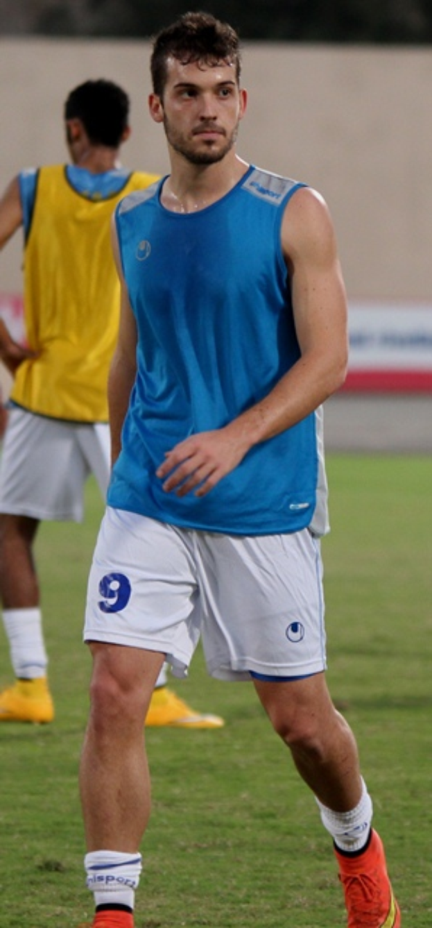 Ein Mann in einem blauen T-Shirt und weißen Shorts steht auf einem Fußballfeld, trägt Fußballschuhe und hat eine Wand und Bäume im Hintergrund.