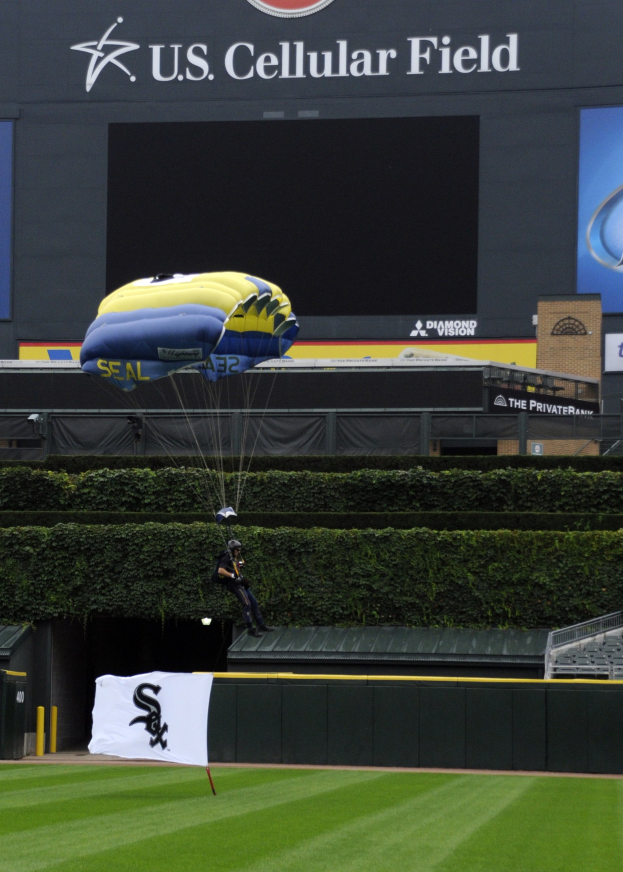 Eine Person, die über einem Baseballfeld mit einem Fallschirm paragleitet, umgeben von Grünfläche, einem Banner, Tafeln mit Text und Logos und einem Gebäude im Hintergrund.