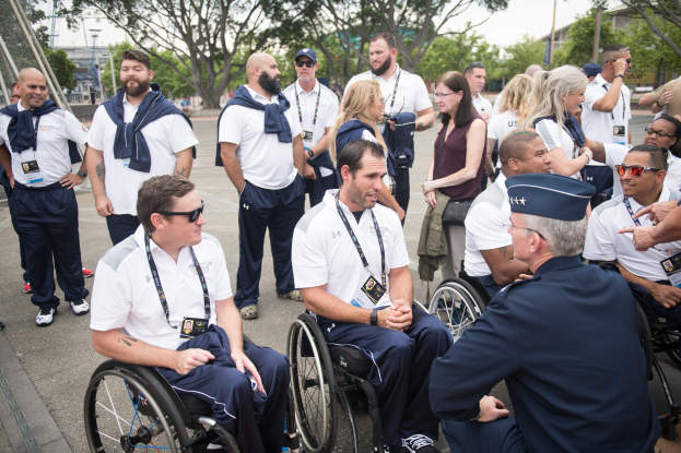 Eine Gruppe von Menschen im Rollstuhl vor einem Hintergrund aus Bäumen, Pfählen, Gebäuden und einem klaren blauen Himmel, wobei einige Mützen und Schutzbrillen tragen, was darauf hinweist, dass sie bei den Paralympischen Spielen sind.