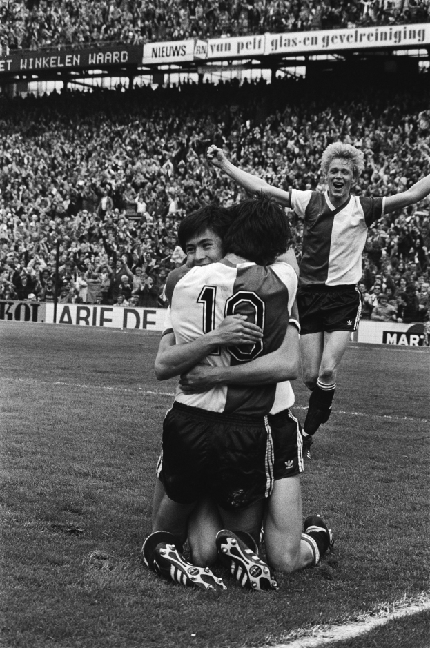 Drei Fußballspieler, die sich auf dem Boden nach einem Torjubel umarmen, mit einer jubelnden Menge und schwenkenden Fahnen im Hintergrund, in einer schwarz-weißen Stadionatmosphäre.
